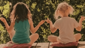 Two female children with their backs to the screen sitting cross legged in front of a a green bush